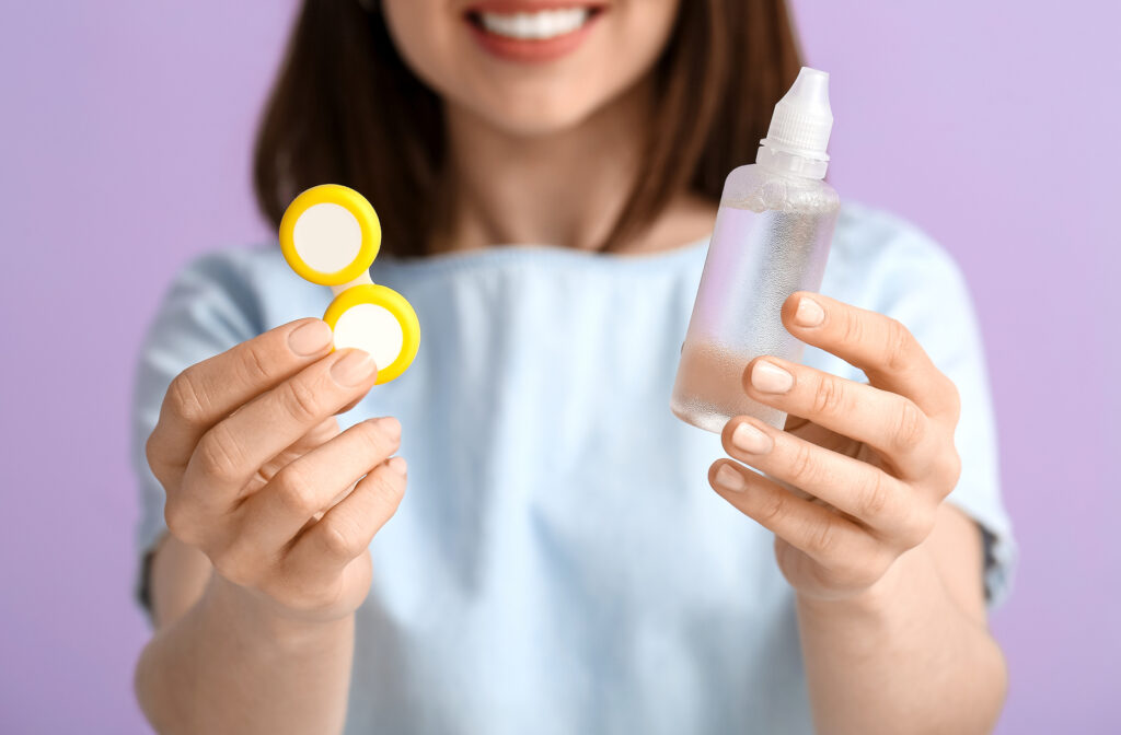 A woman photographed from the mouth down smiles at camera from background, and holds a yellow contact lens case and solution in foreground