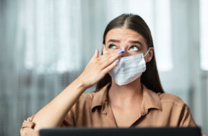 A young woman wearing a surgical mask with allergies is touching is itchy eyes due to dry eyes.