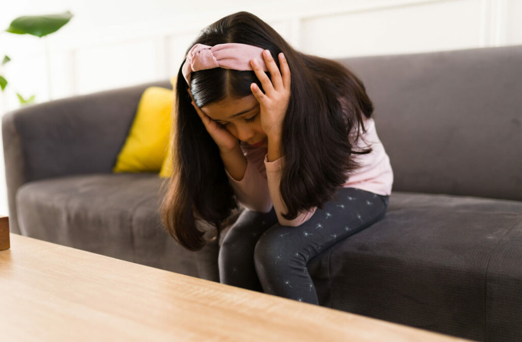 A young girl with long hair is touching her head with both hands while sitting on a sofa and is suffering from a headache.