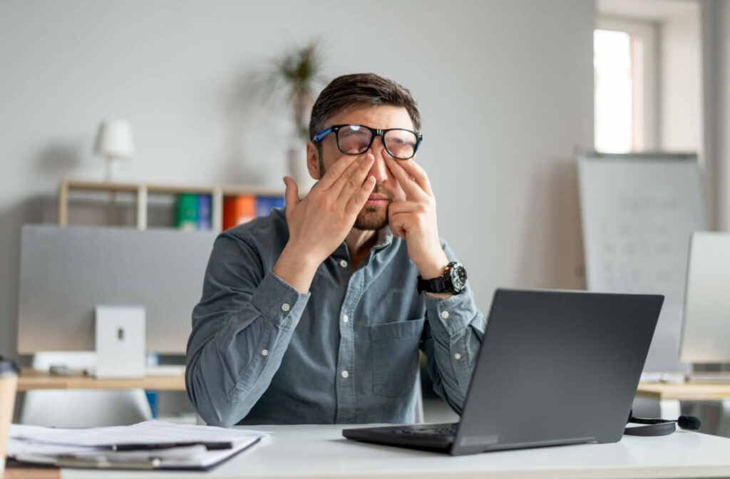 A professional looking young man rubs his eyes with both of hands due to stress and dry eyes in front of his laptop.