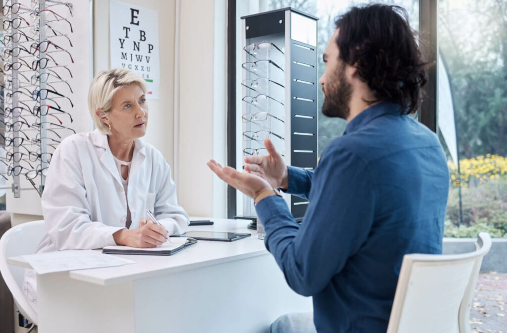 A man in a blue shirt sharing details of his droopy eyelids to his optometrist who is listening diligently while taking notes.
