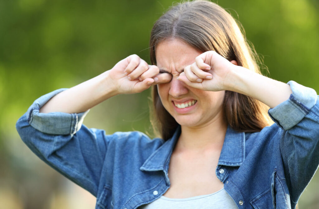 A young woman standing outside and rubbing her eyes with both hands, making a face as if in pain
